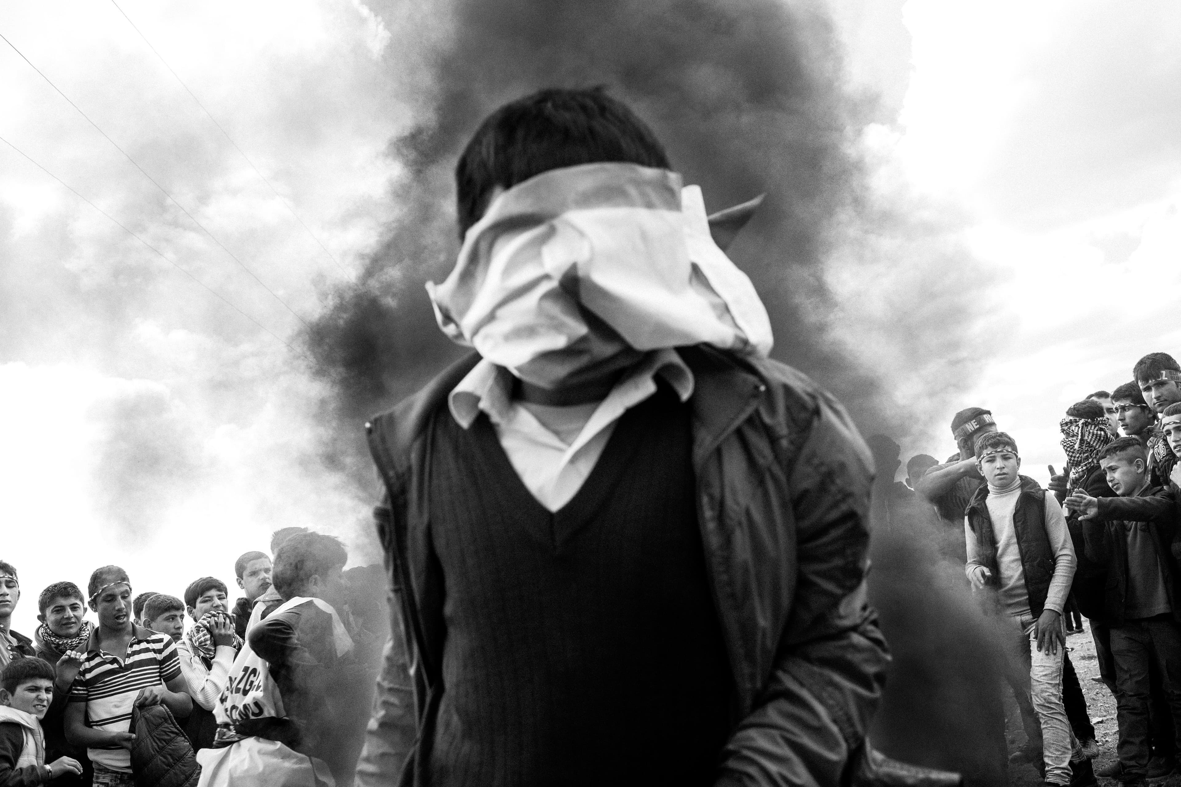 Images by Turkish photographer Furkan Temir documenting the war in Syria. This image is of a child with cloth covering the entirety of his face. Behind him stands a plume of smoke and a wall of other Syrian refugees.